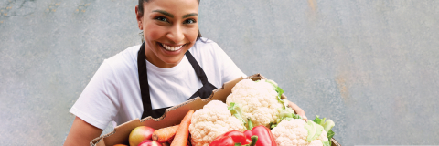 This banner image shows a woman holding a fresh vegetable box and smiling while getting her picture taken.
