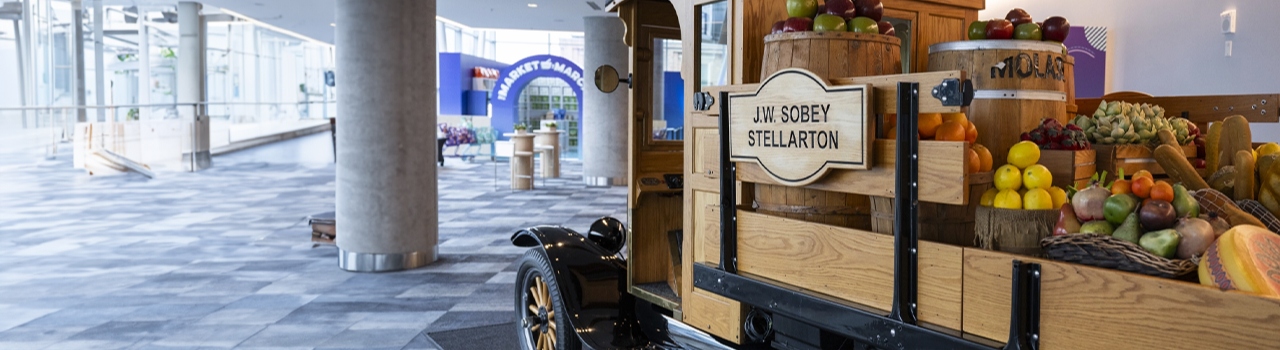 A vintage truck interior display loaded with an assortment of fruits and vegetables is seen in a modern, brightly lit indoor space with large windows and columns. The truck has a wooden sign that reads "J.W. Sobey Stellarton.