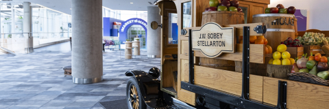 A vintage truck interior display loaded with an assortment of fruits and vegetables is seen in a modern, brightly lit indoor space with large windows and columns. The truck has a wooden sign that reads "J.W. Sobey Stellarton.