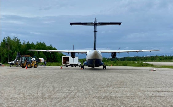 A small plane is parked on a tarmac against a backdrop of cloudy skies and green trees. Orange cones are placed around the area, suggesting it is an active work zone.