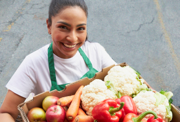 This banner image shows a woman holding a box of fresh vegetables and smiling while her picture is being taken.