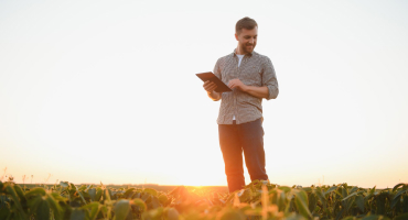 men are standing in a green crop field under a clear sky. One man is showing something on a tablet to the other man.
