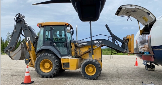 An images shows a front loader is transferring cargo into an airplane's open rear compartment on a tarmac. Orange traffic cones are placed near the machinery and the aircraft.