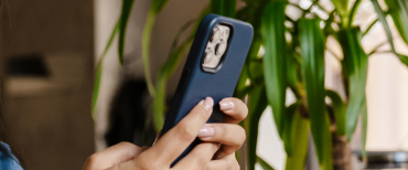 A person is holding a smartphone with a blue case, standing next to a green potted plant.